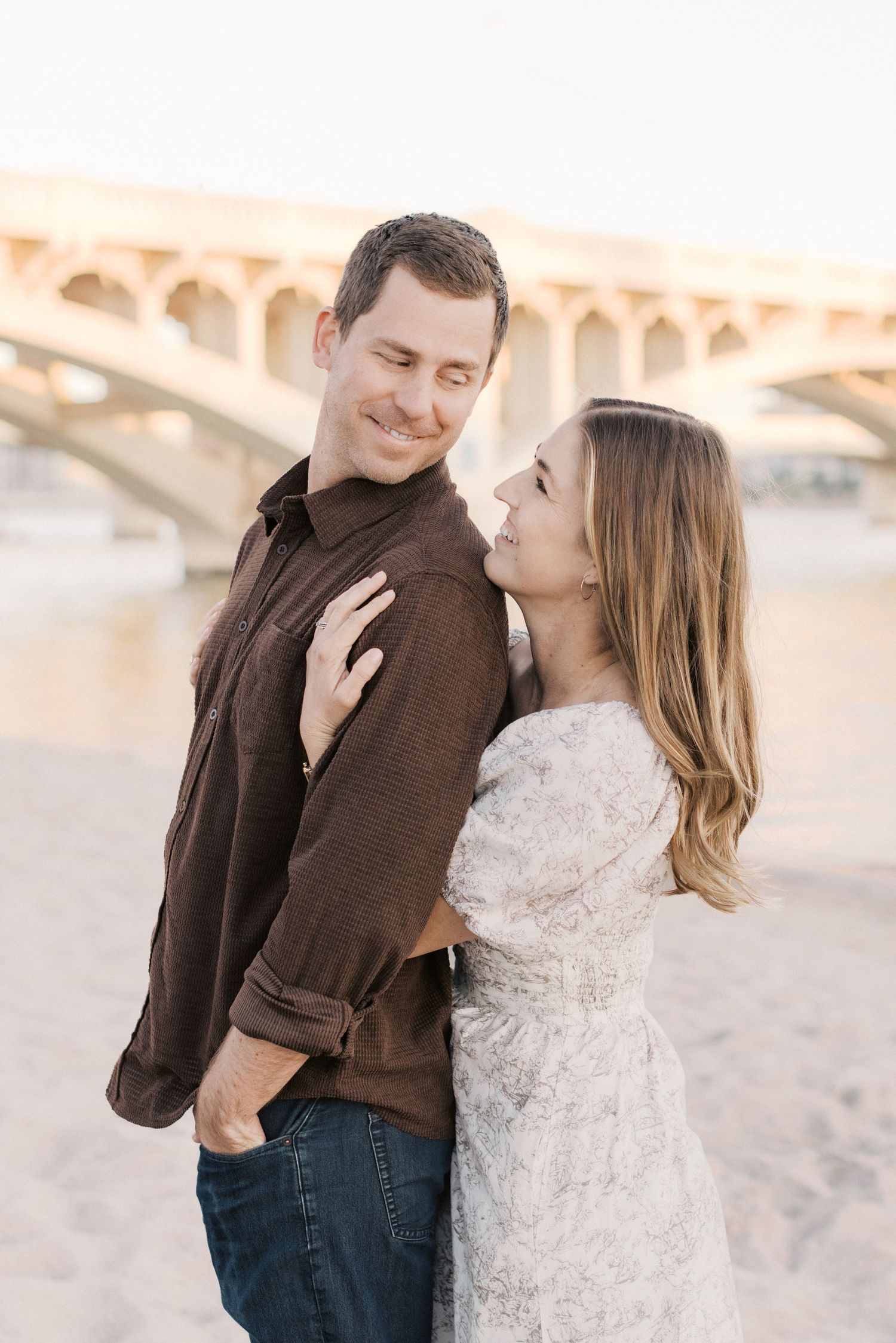 Couple embracing on sandy beach near bridge at sunset, woman in floral dress smiling up at man in brown shirt.