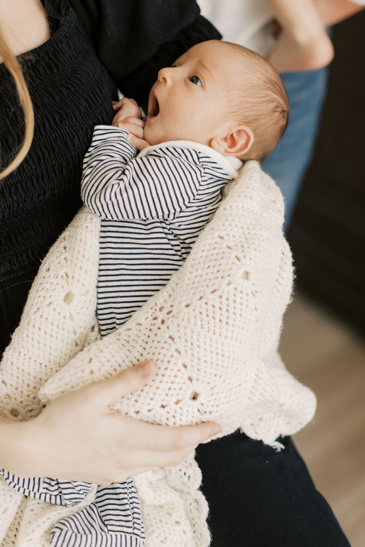A newborn baby in striped onesie wrapped in cream knit blanket being cradled in an adult's arms.