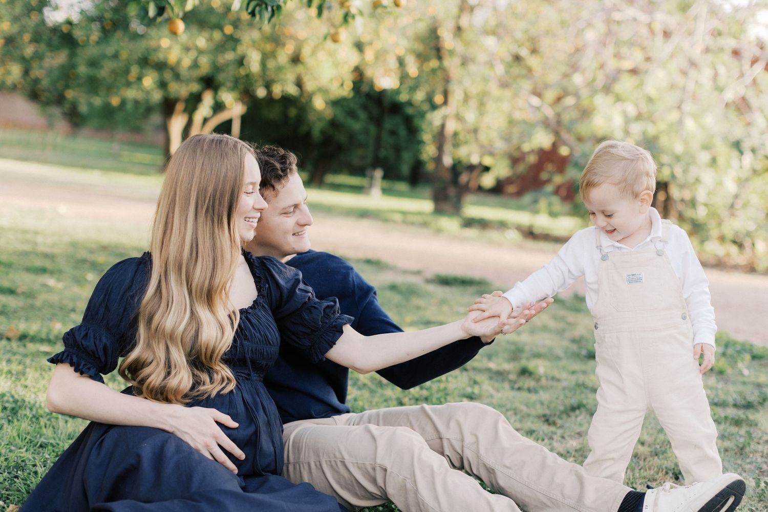 Family of three sitting on grass in a park, parents smiling at toddler in cream overalls reaching out to them.
