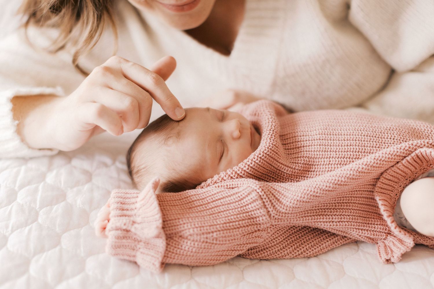 A tender mother gently touches her newborn baby dressed in a pink knit sweater lying on a white quilted surface.