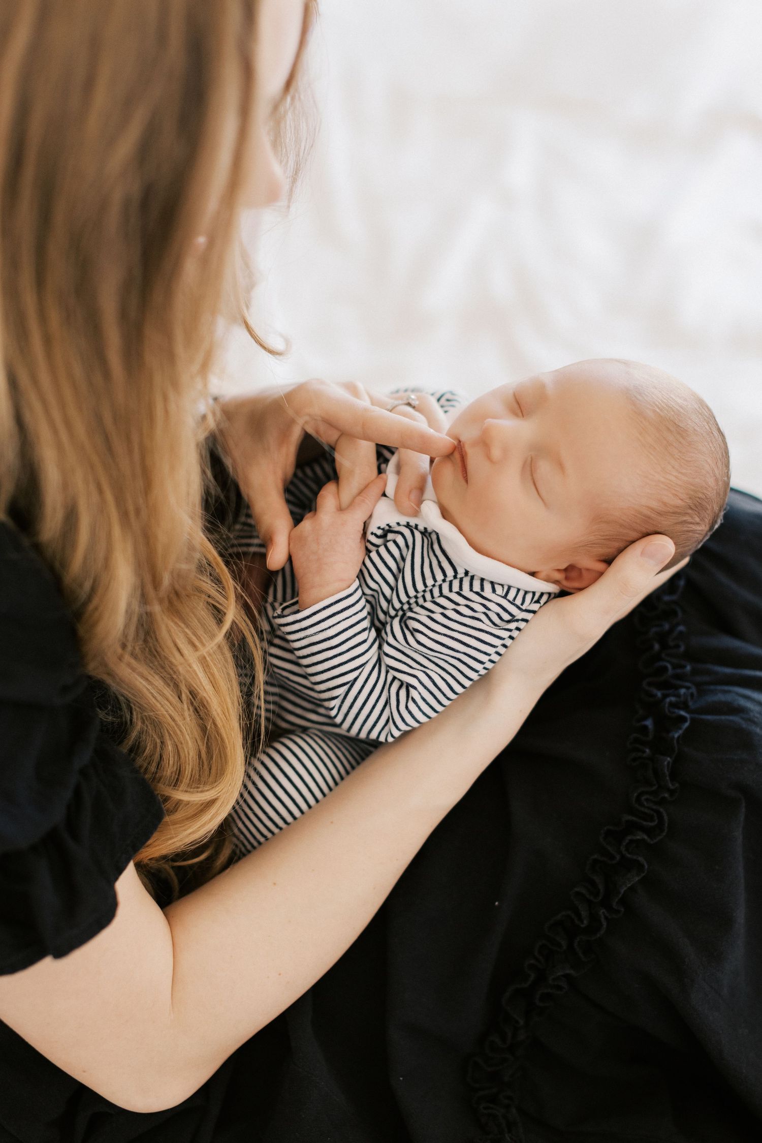 A mother cradles her sleeping newborn baby dressed in a striped onesie, gently touching the infant's tiny hand.