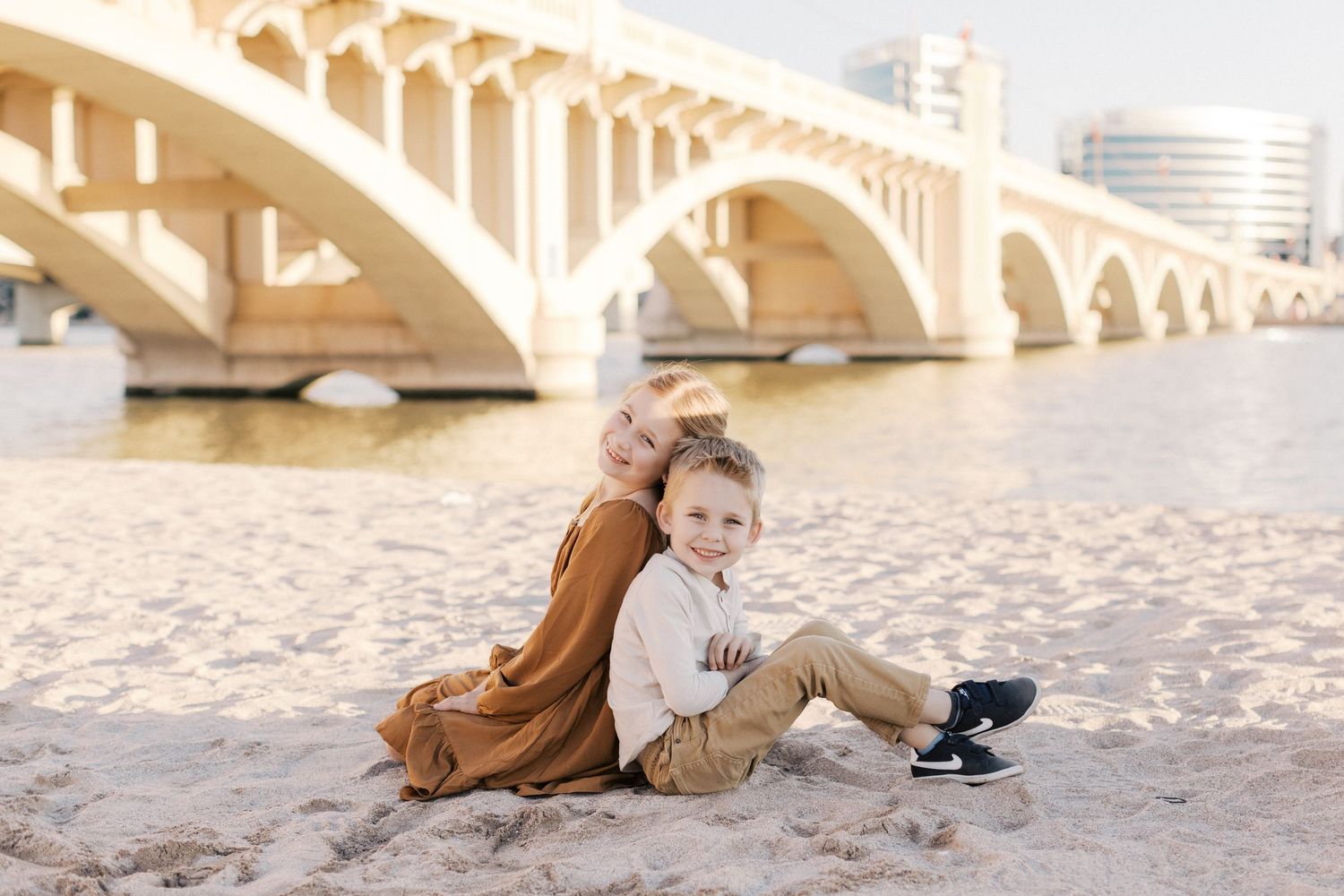 A mother and young boy sit together on a sandy riverbank with an arched bridge in the background.