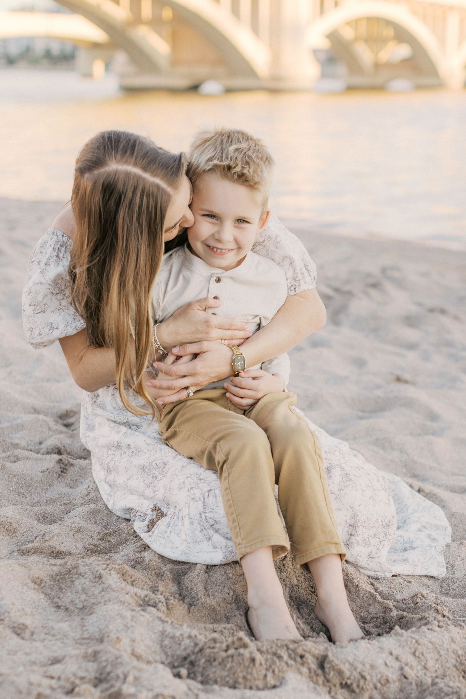 A mother hugs her smiling young son on a sandy beach near a bridge at sunset.