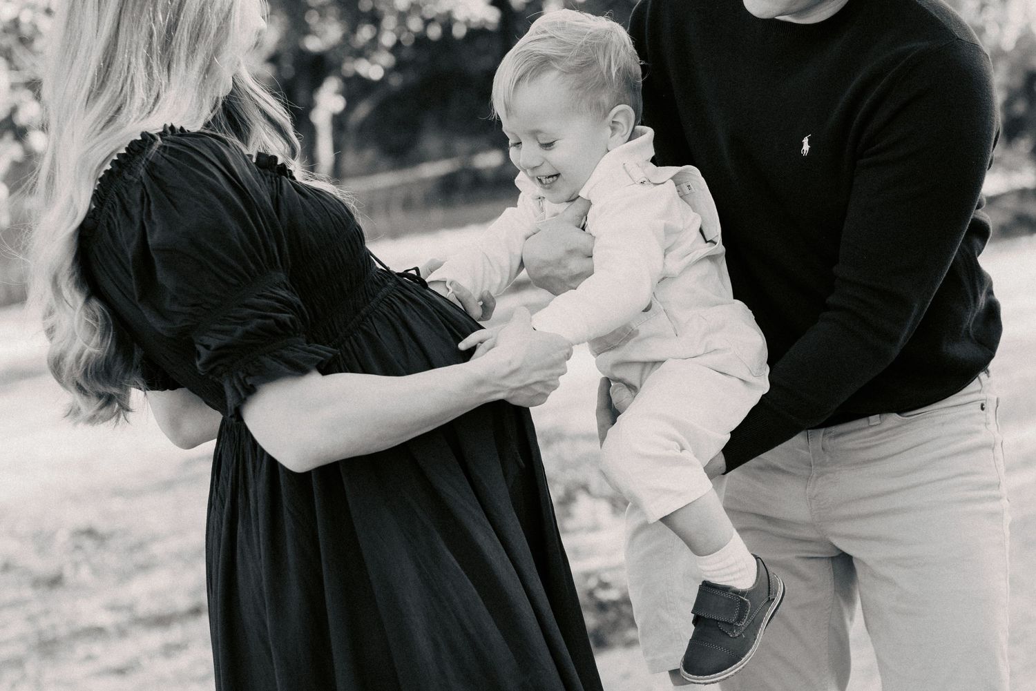 Black and white family photo of parents with a laughing baby outdoors in a park setting.