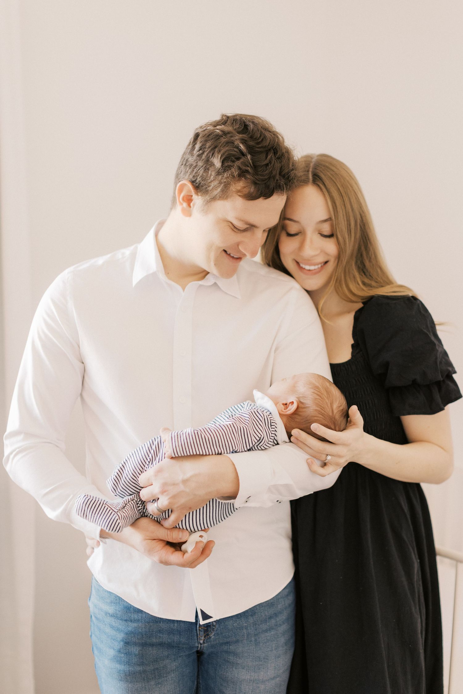 A young couple smiles lovingly at their newborn baby, the father in a white shirt and mother in a black dress.