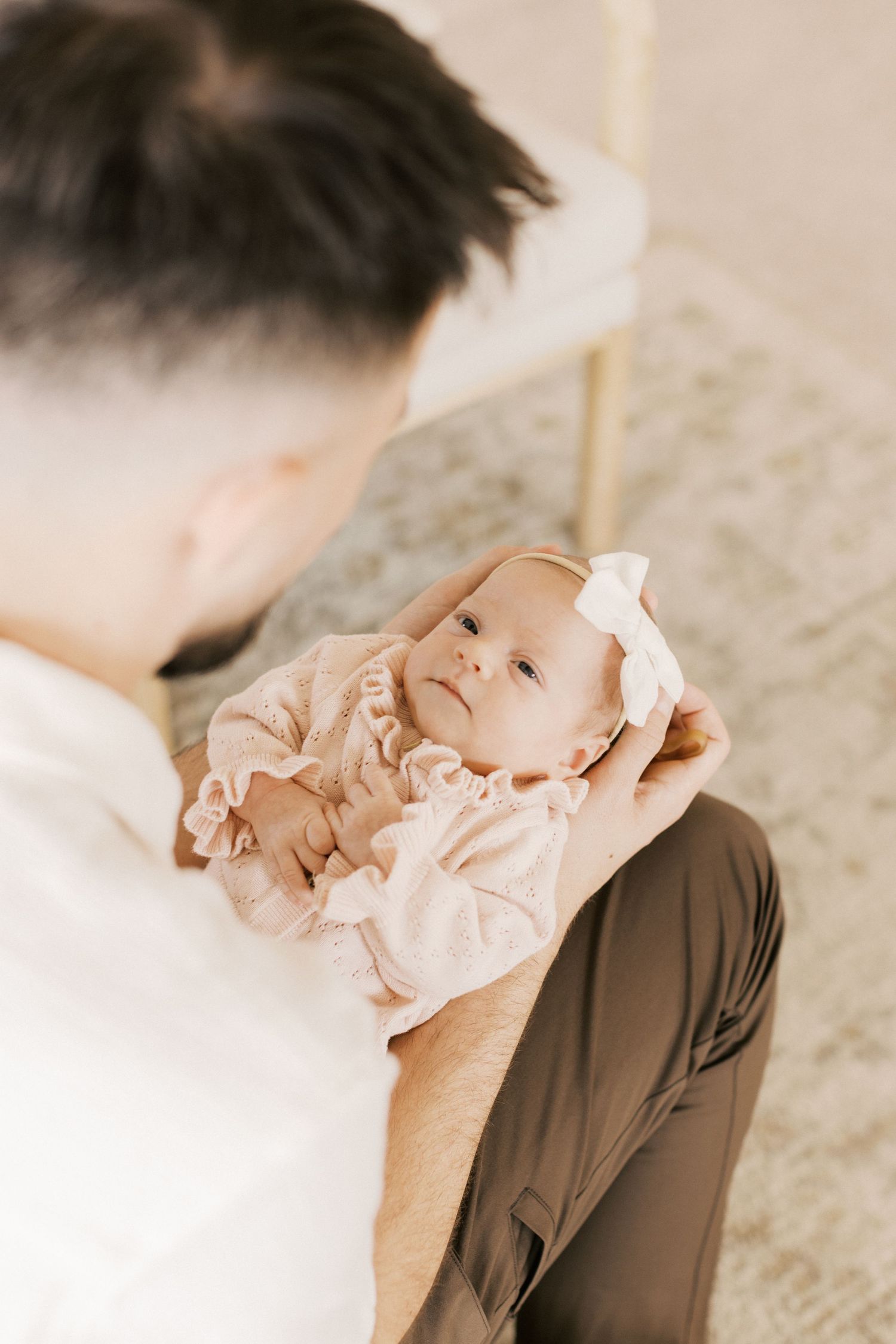 A father cradles his newborn baby girl dressed in a pink outfit with a bow headband during a lifestyle newborn session.
