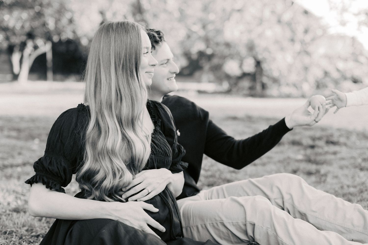 Black and white photo of a pregnant couple sitting on grass in a park, woman with long hair, man pointing.