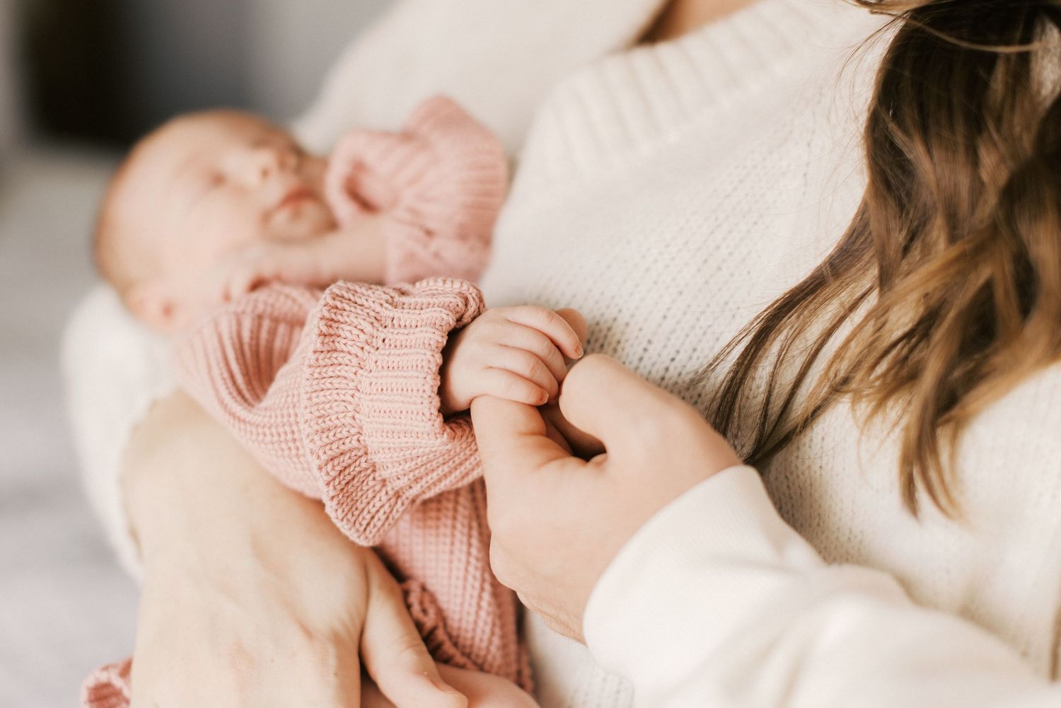 Mother in white knit sweater cradling newborn baby dressed in pink knit outfit, gently holding tiny hands.