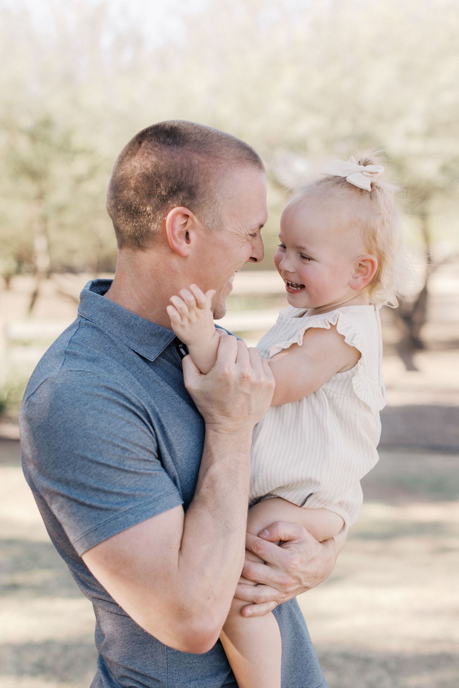Loving father holding laughing baby girl outdoors, sharing a joyful nose-to-nose moment in sunlit park.