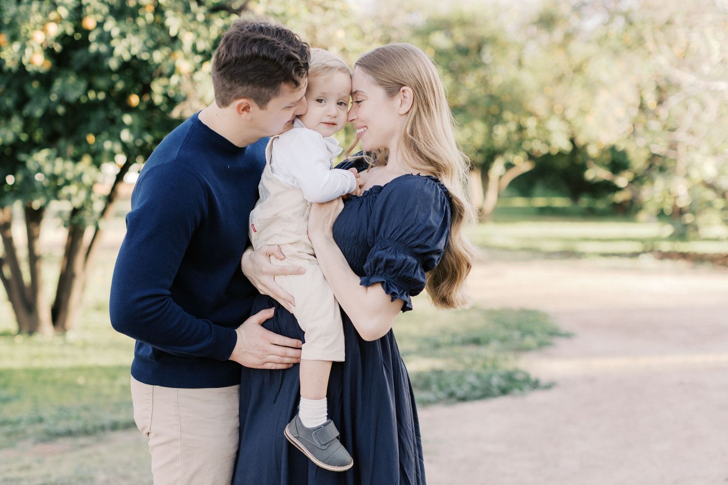 Young family with a toddler sharing a tender moment outdoors in a sunlit park, dressed in navy and neutral tones.