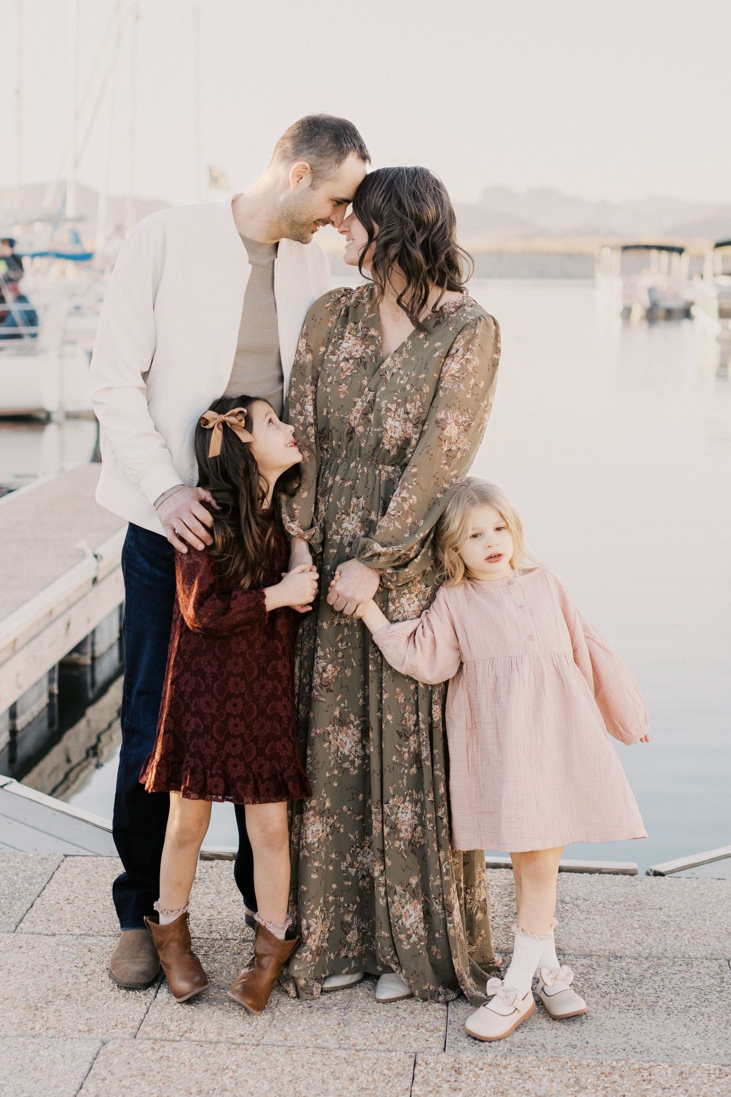 Romantic family portrait of couple with two daughters embracing near marina waterfront at golden hour.