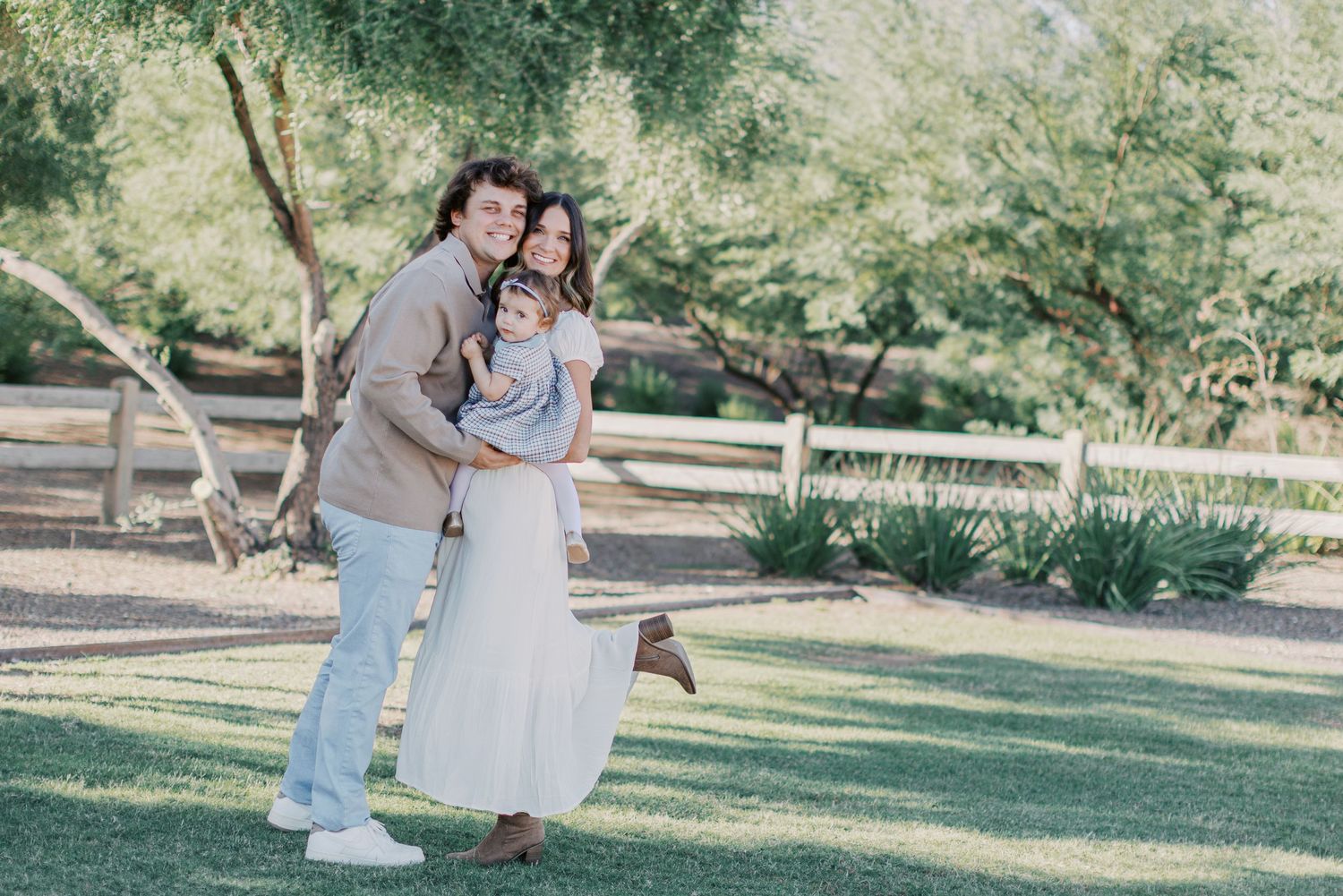 A happy family of three poses outdoors on green grass near a white fence surrounded by lush trees.