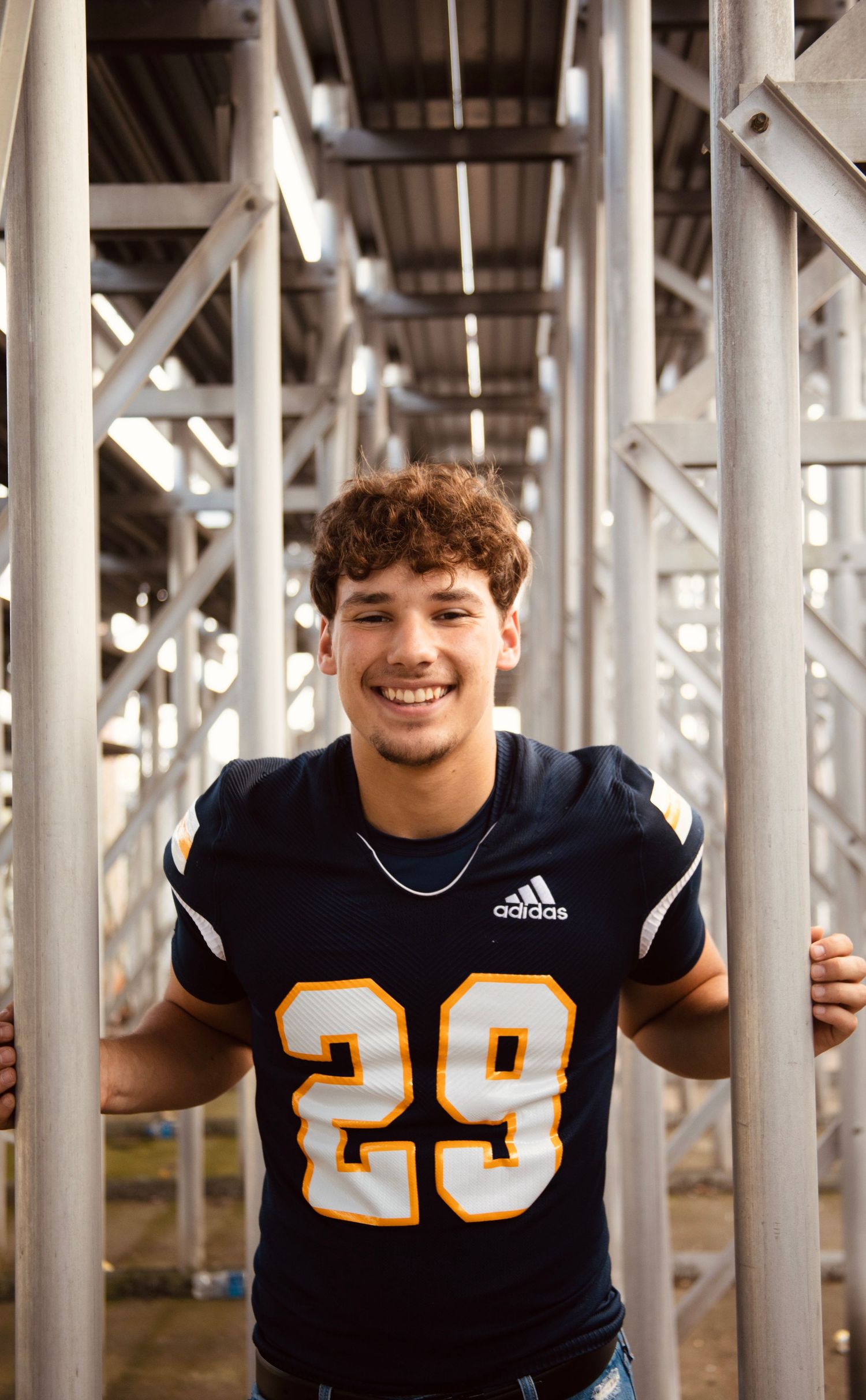 Three football players in navy jerseys with gold numbers pose under stadium bleachers.