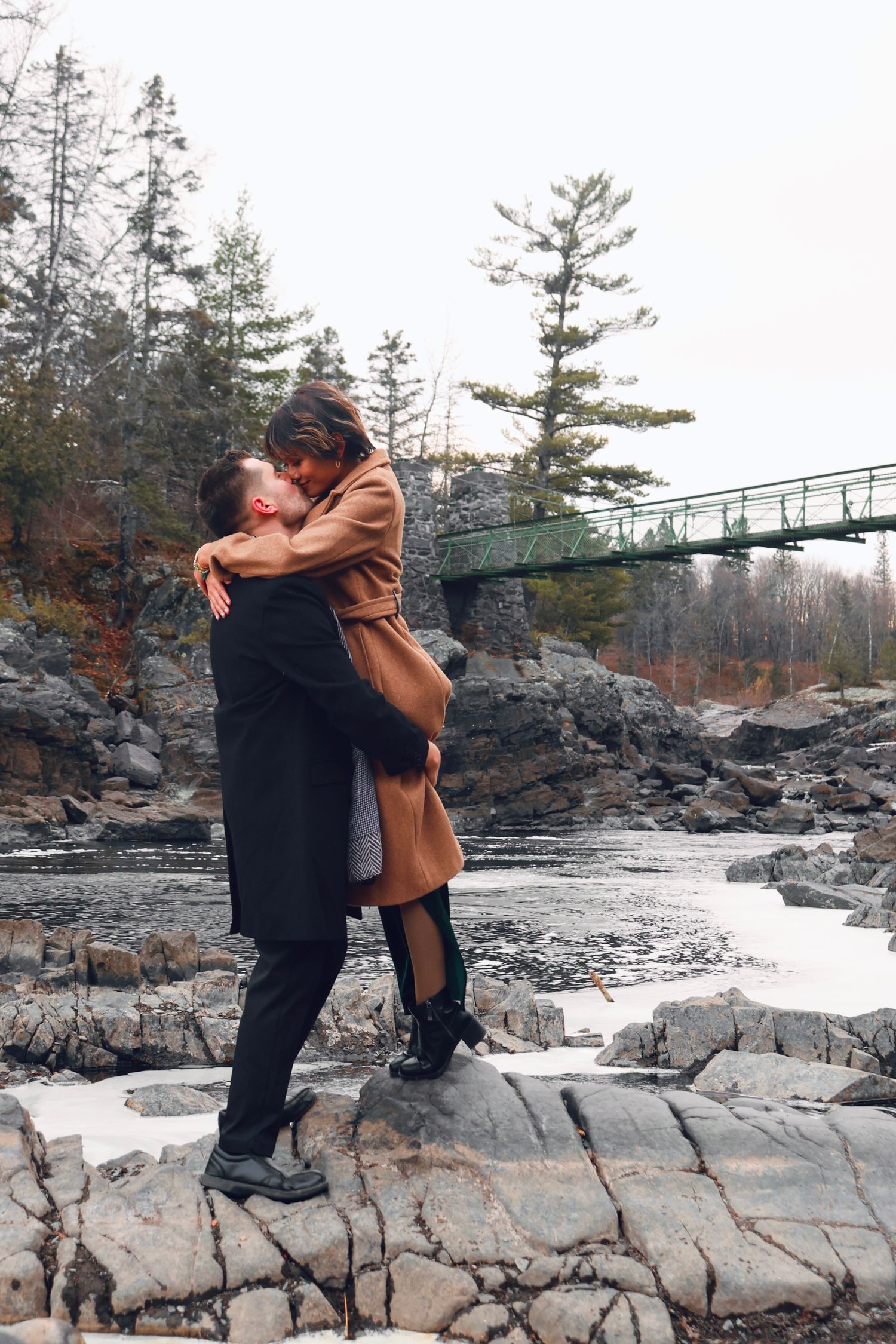 Two people embrace romantically on rocky coastal terrain with evergreen trees and a footbridge in the background.