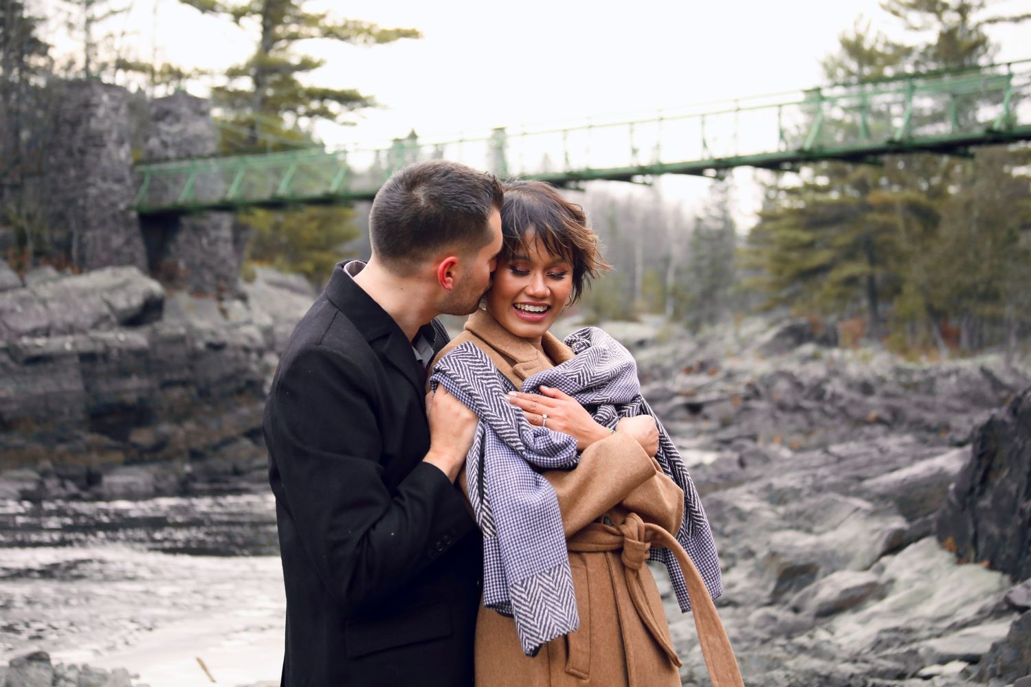 A couple embraces on rocky terrain with evergreen trees in the background.