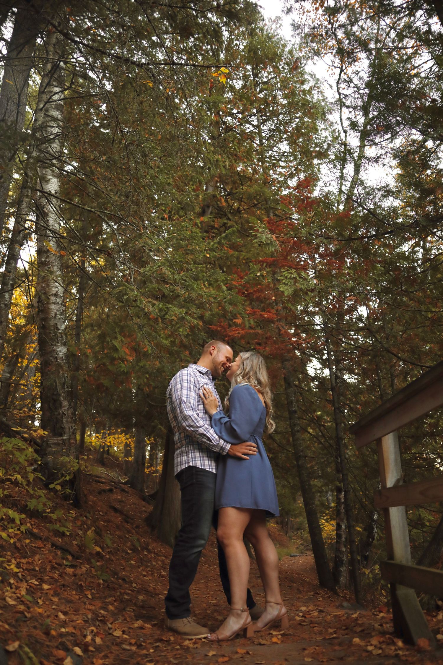 Two people share an intimate moment on a wooded trail surrounded by fall foliage.
