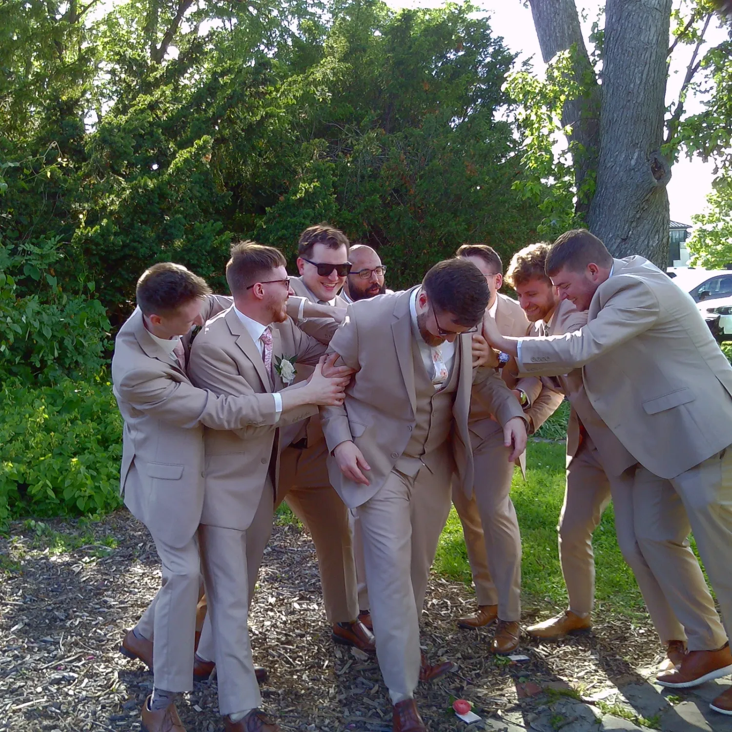 Wedding party in tan suits poses playfully outdoors under trees.