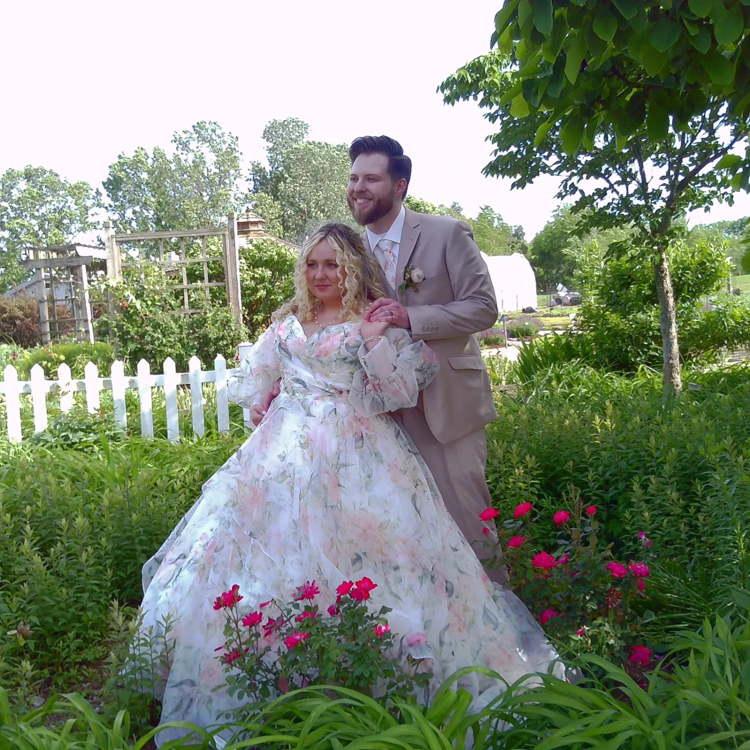 A couple in a garden setting with pink roses and white picket fence share a romantic dance-like moment in formal attire.