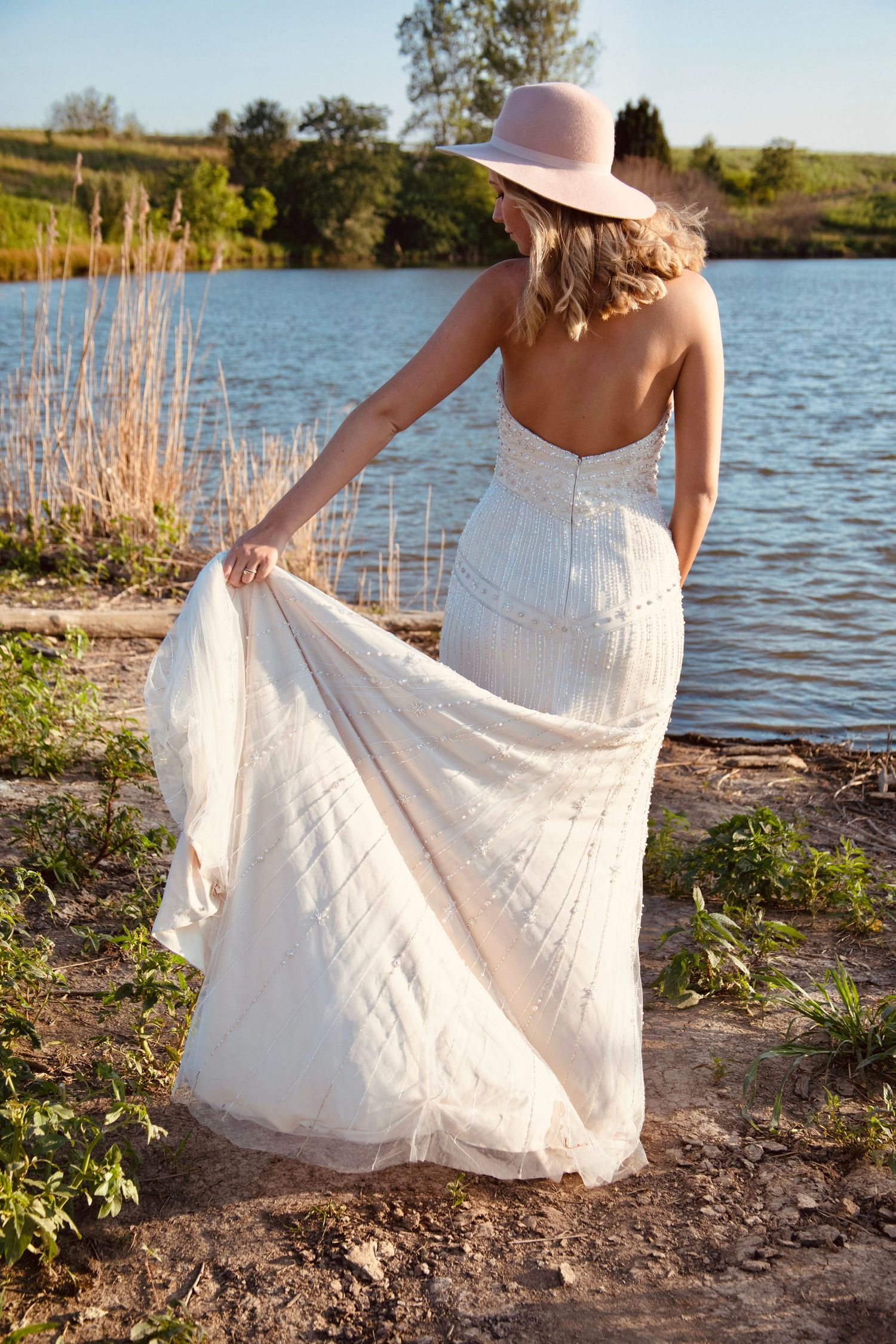 A person in a flowing white dress and pink hat twirls gracefully by a serene lakeside during golden hour.
