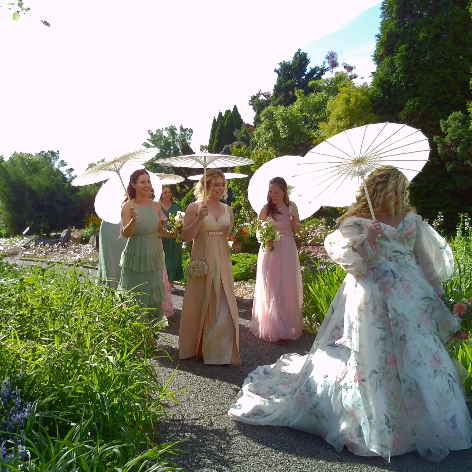 A group in pastel dresses and parasols walk along a garden path in an elegant outdoor setting.