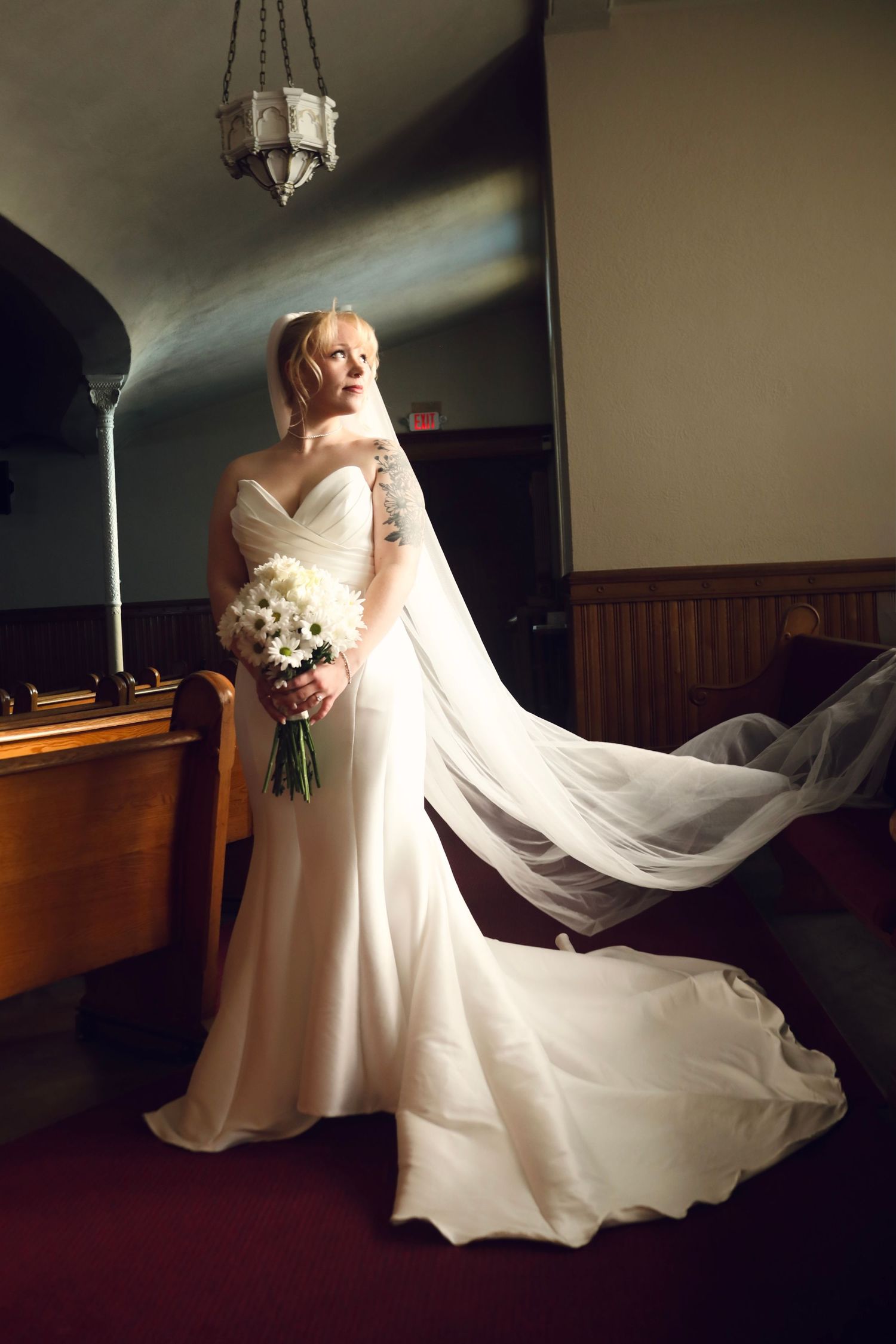 Elegant wedding dress with flowing train and white bouquet photographed in a church setting with vintage chandelier.