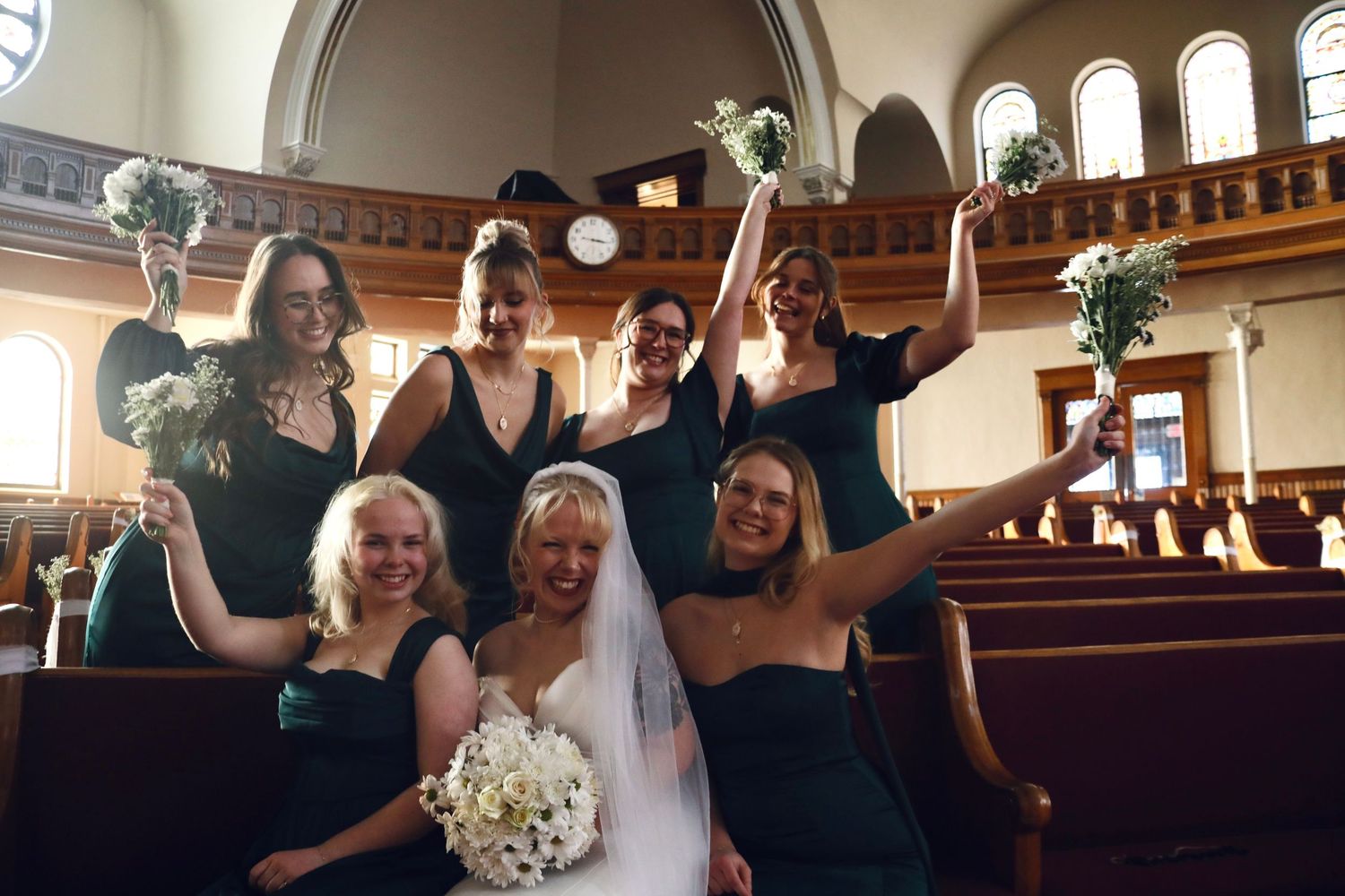 Wedding party in navy dresses celebrating in church pews with bouquets raised high.