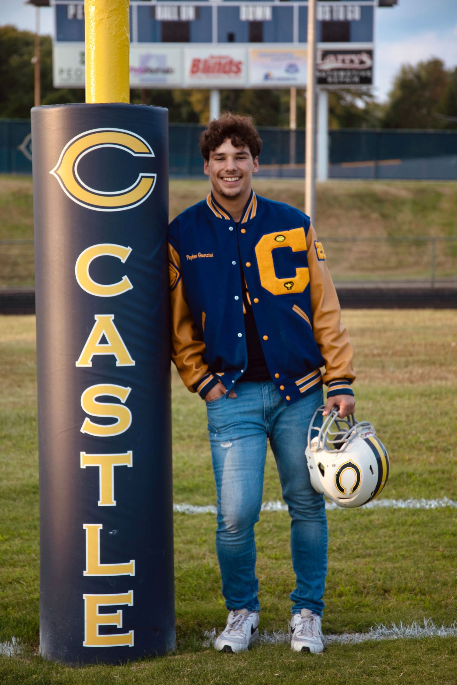 A football player poses with a ball next to blue and gold lettered goal posts on a grass field.