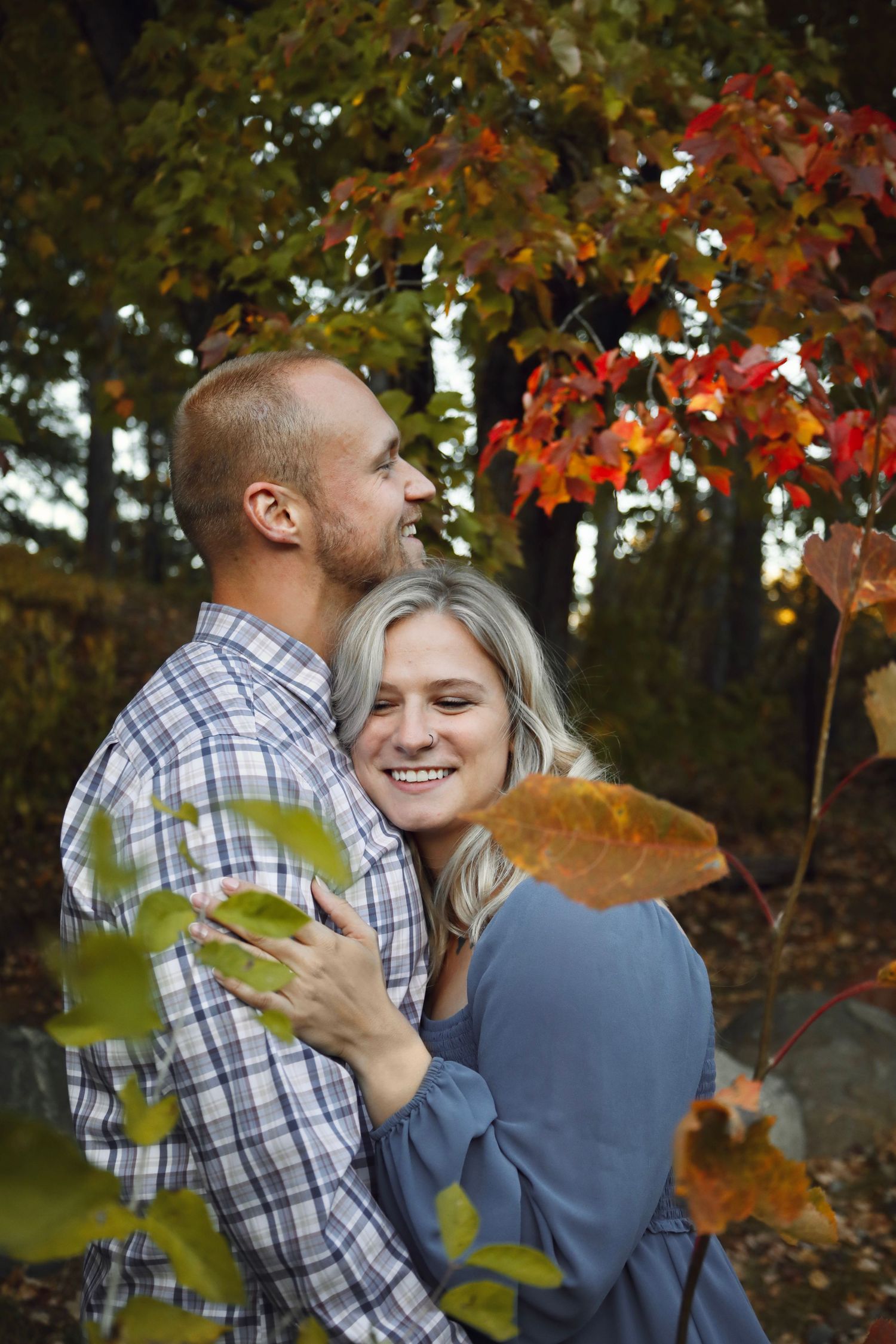 A couple embraces lovingly among autumn trees with orange and red leaves in the background.