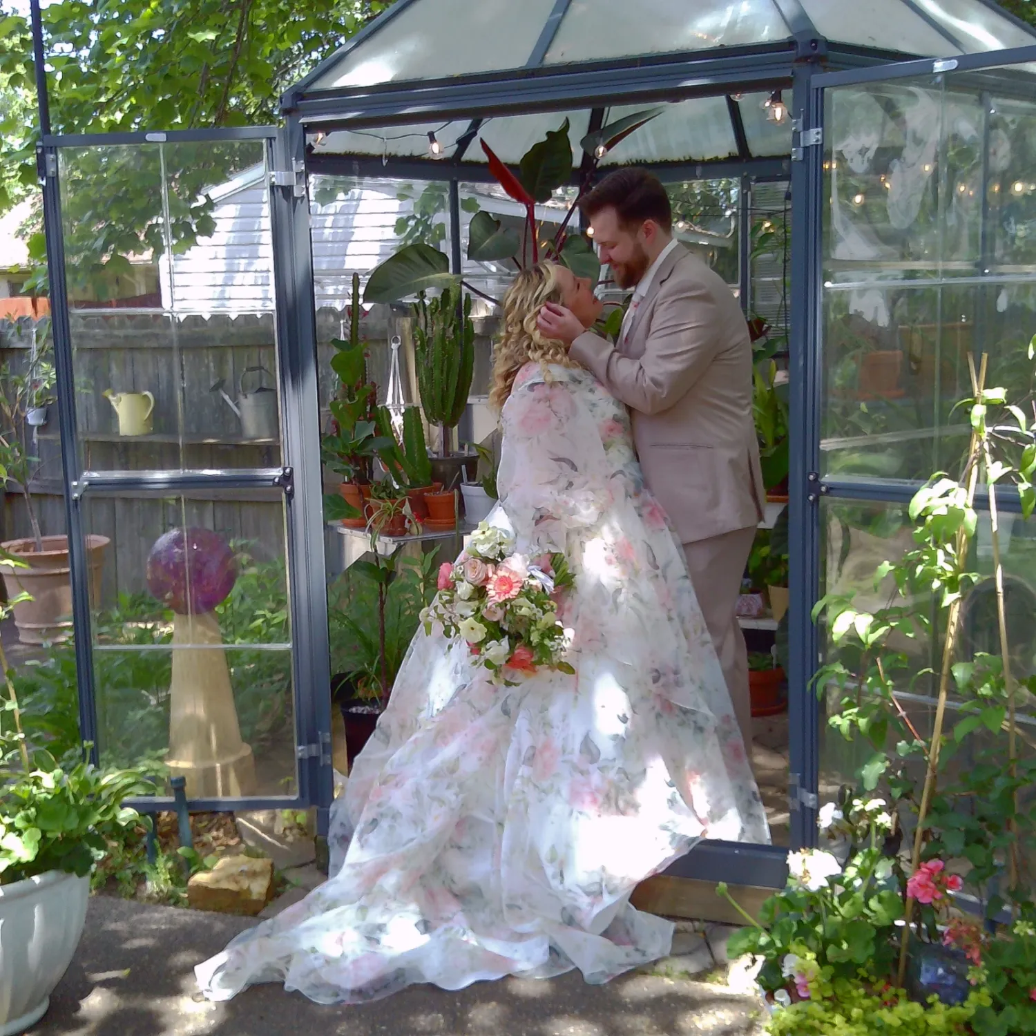 A couple in formal wedding attire poses together in a lush greenhouse setting with natural sunlight streaming in.