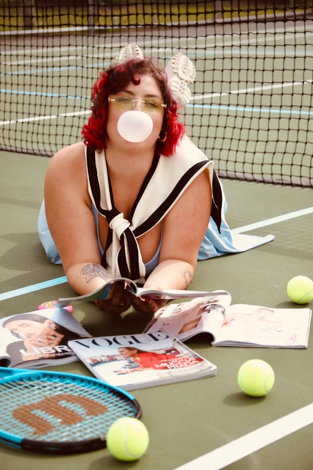 Person lying on tennis court reading magazines with tennis balls and racquet nearby on sunny day.