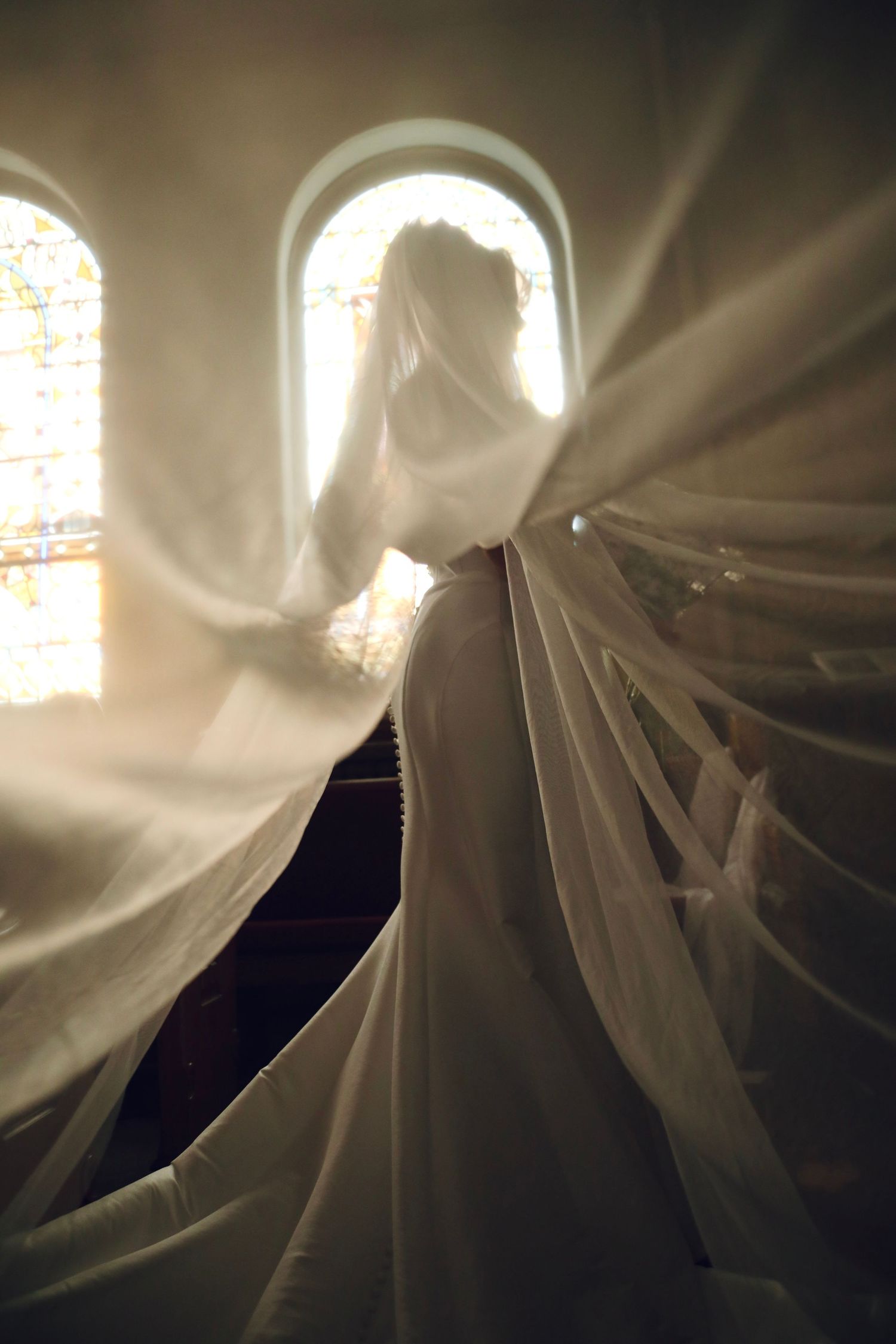Ethereal backlit wedding photo with flowing veil and stained glass windows in church.