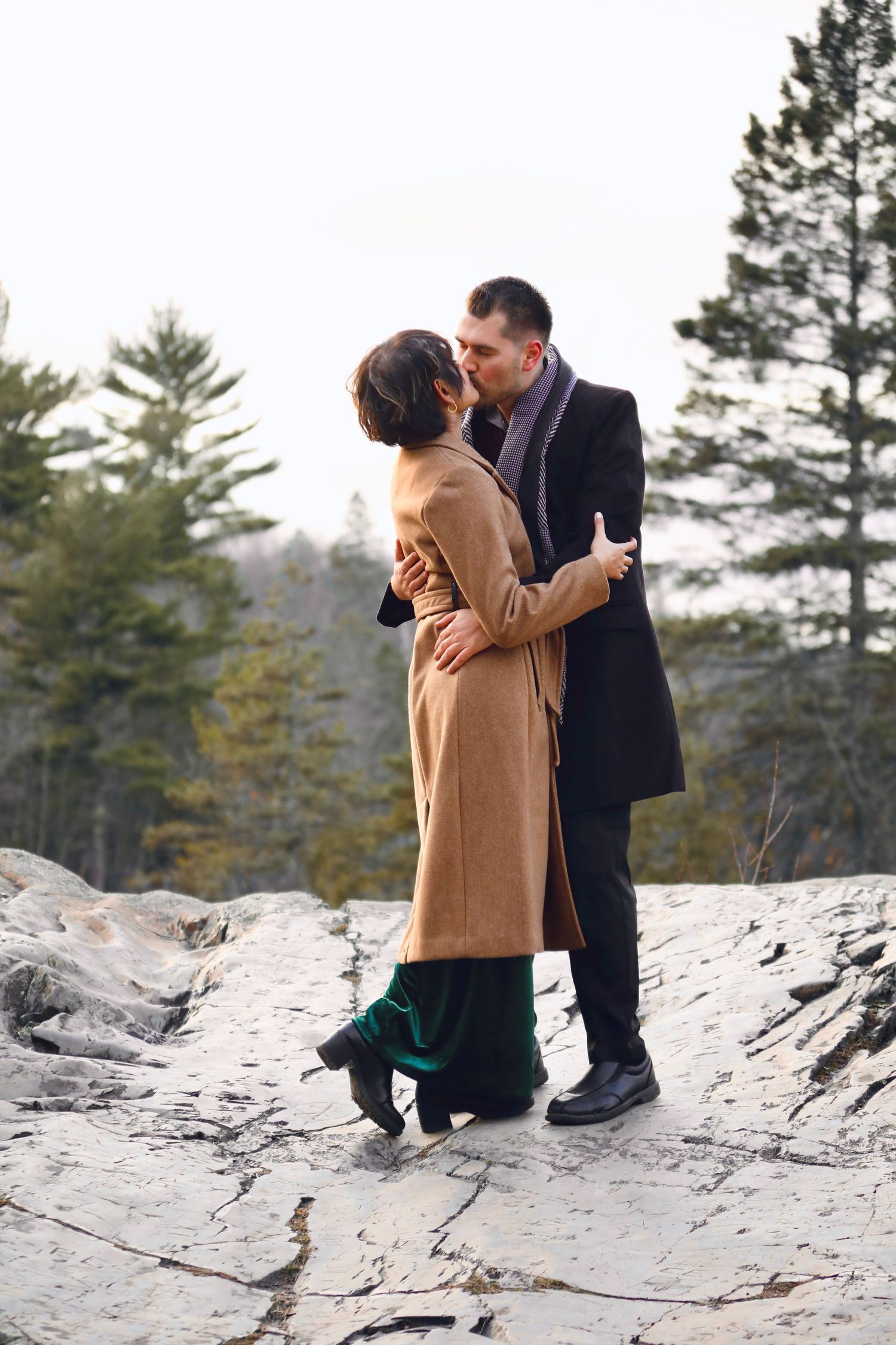 A couple shares an intimate moment on snowy rocks surrounded by evergreen trees.