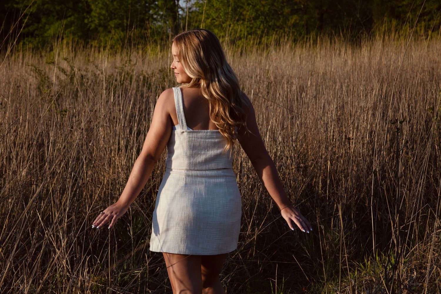 Woman in white dress walking through tall grass field at sunset.