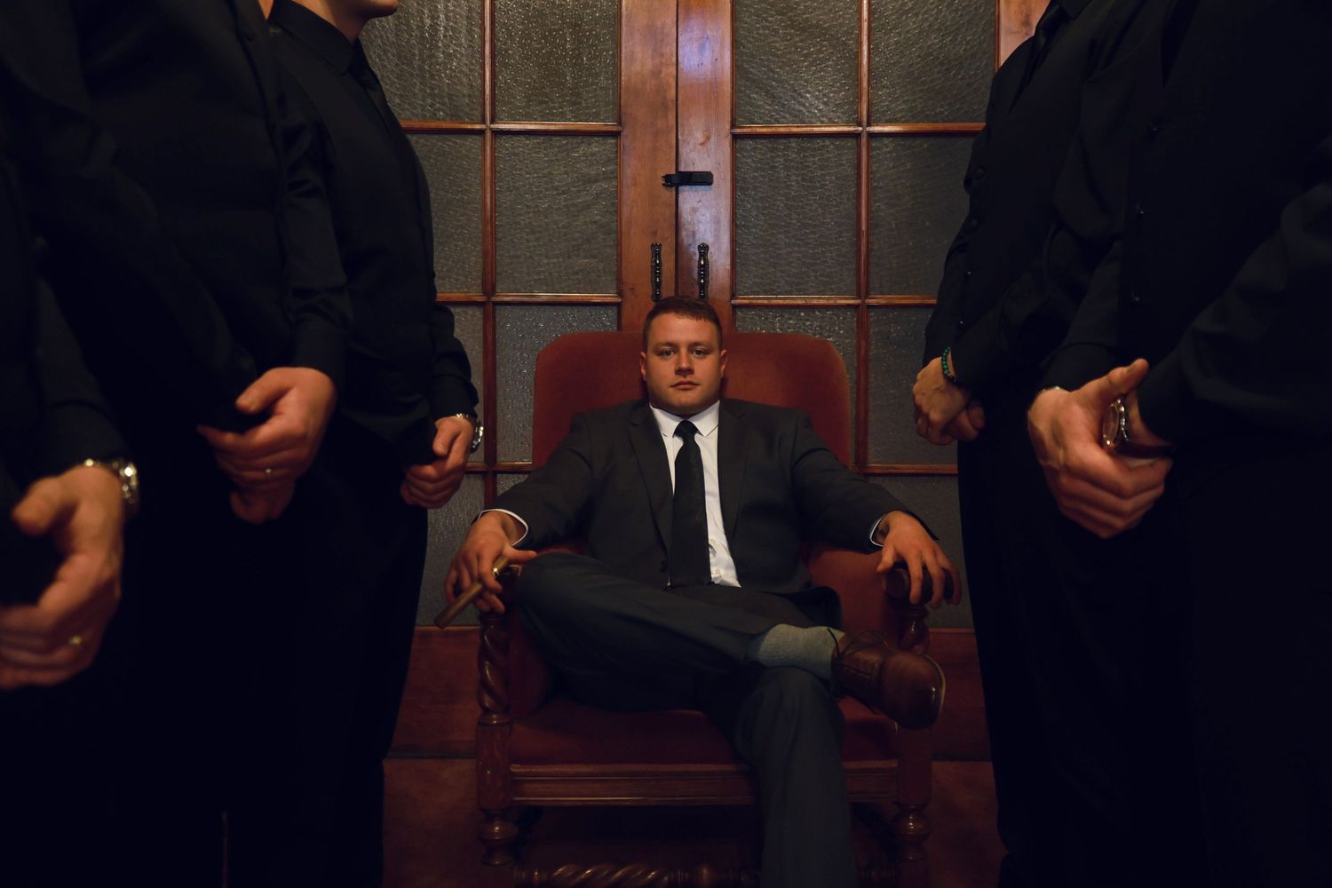 Dramatic portrait of person seated in leather chair flanked by standing figures in dark room.