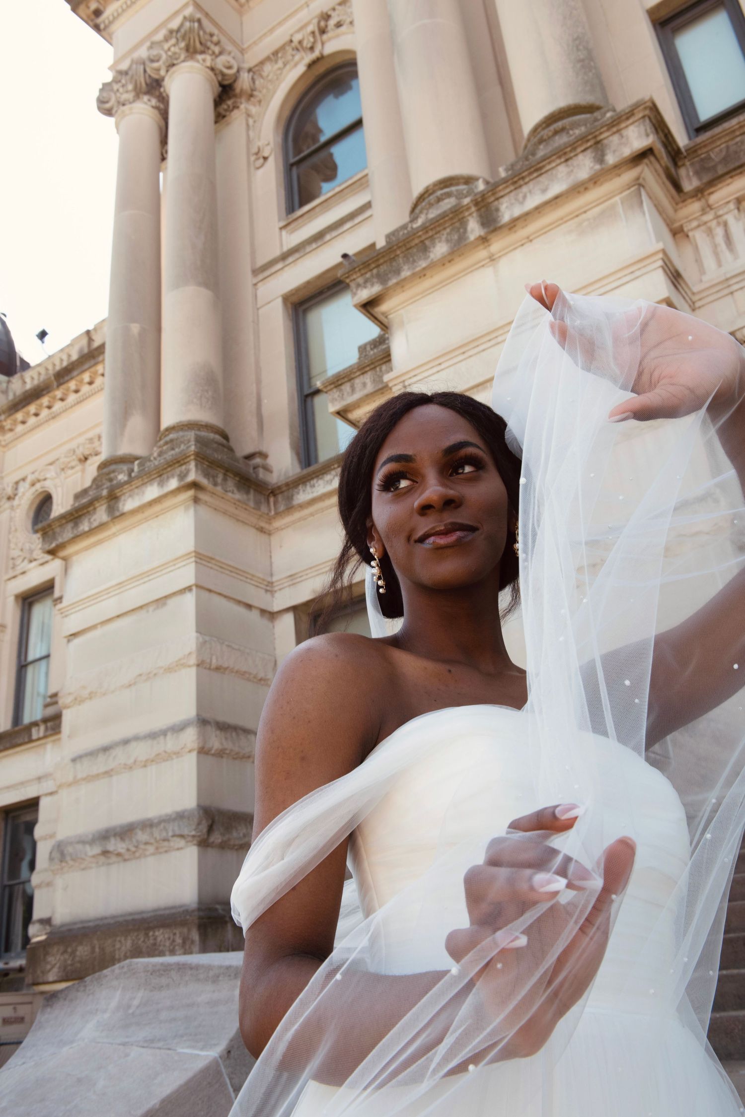 A bride in a flowing white wedding dress with veil poses outside a grand historic building with ornate columns.