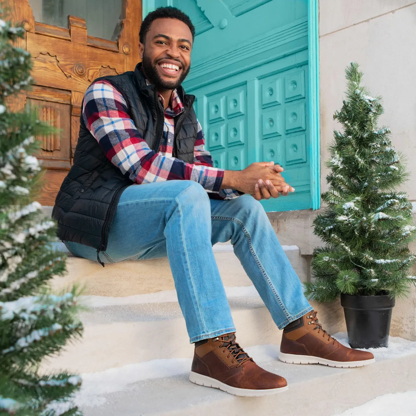 Casual portrait on porch steps with plaid shirt, vest and jeans near decorative evergreens.