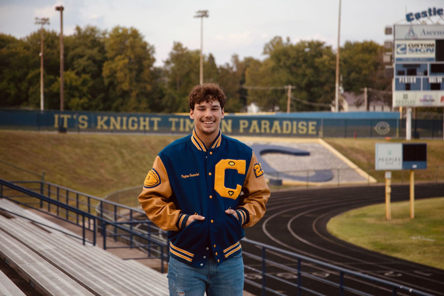 High school athlete in varsity letterman jacket poses on track field during golden hour sunset.