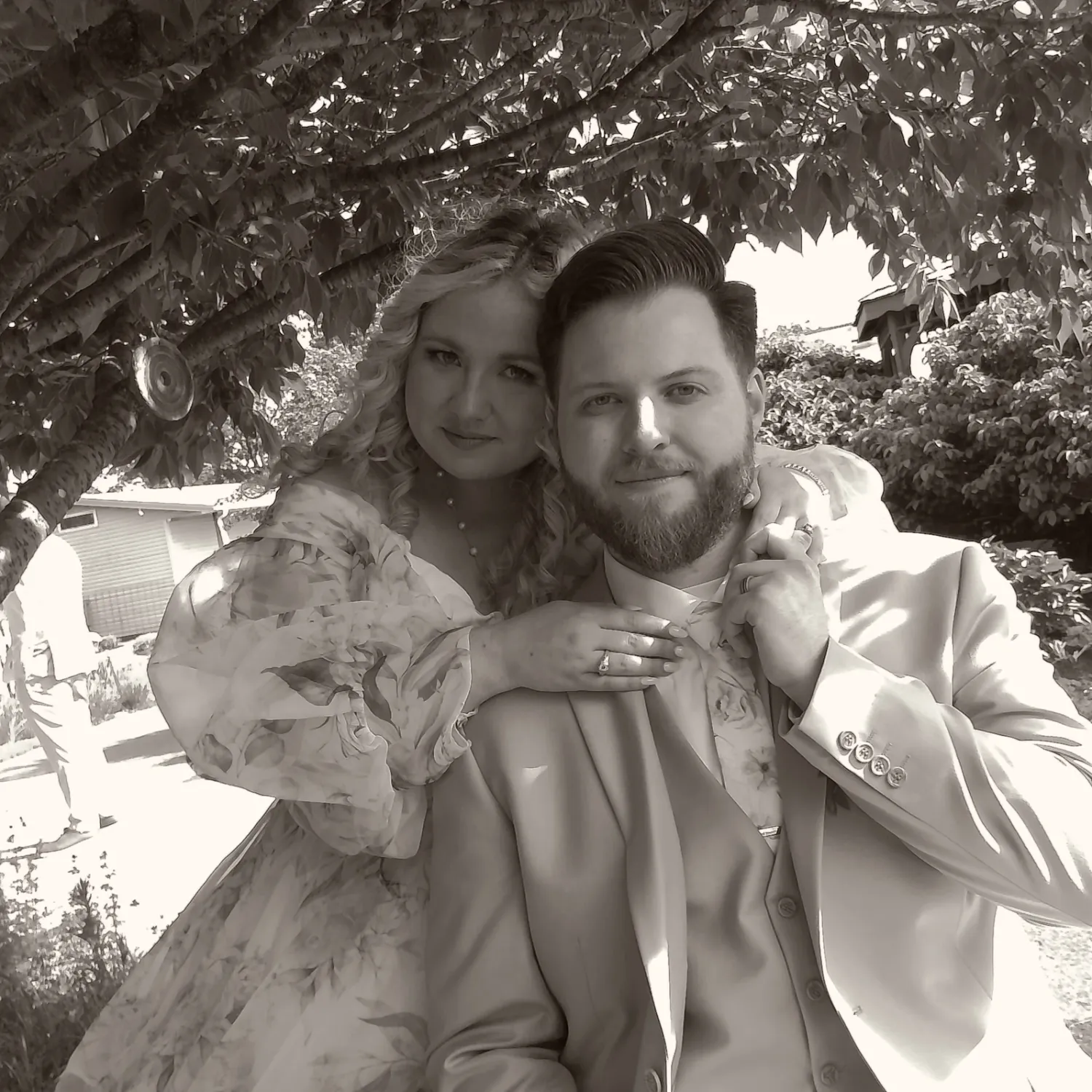 A couple in formal attire pose together outdoors in black and white, with trees and foliage in the background.