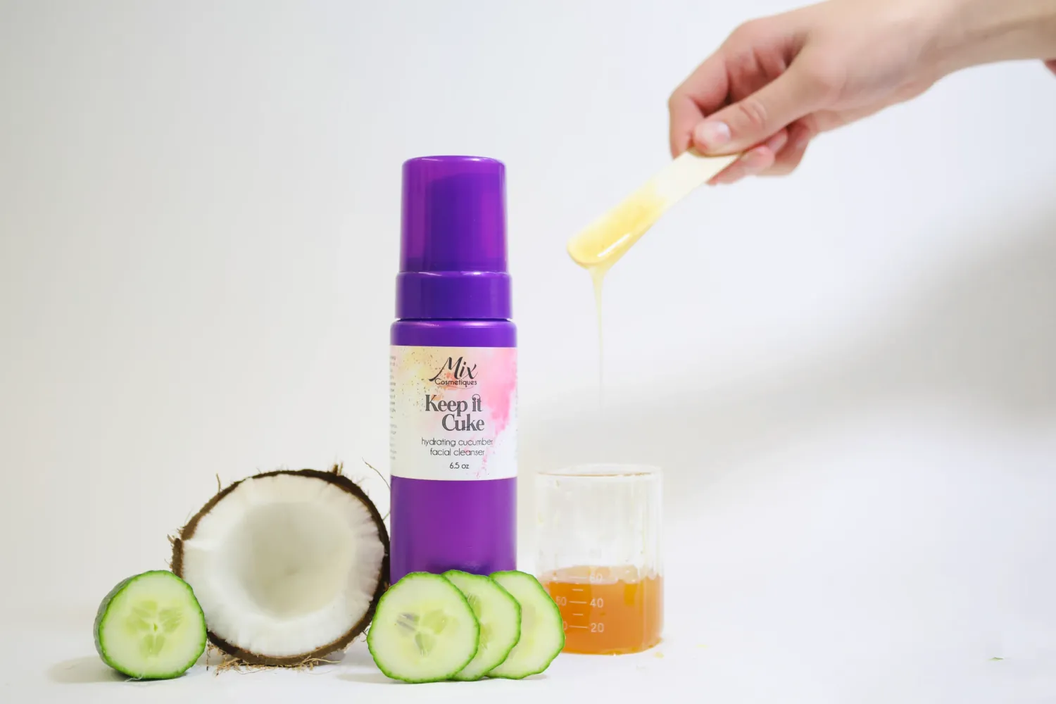Sliced cucumber and coconut next to a purple beauty product bottle on white background.