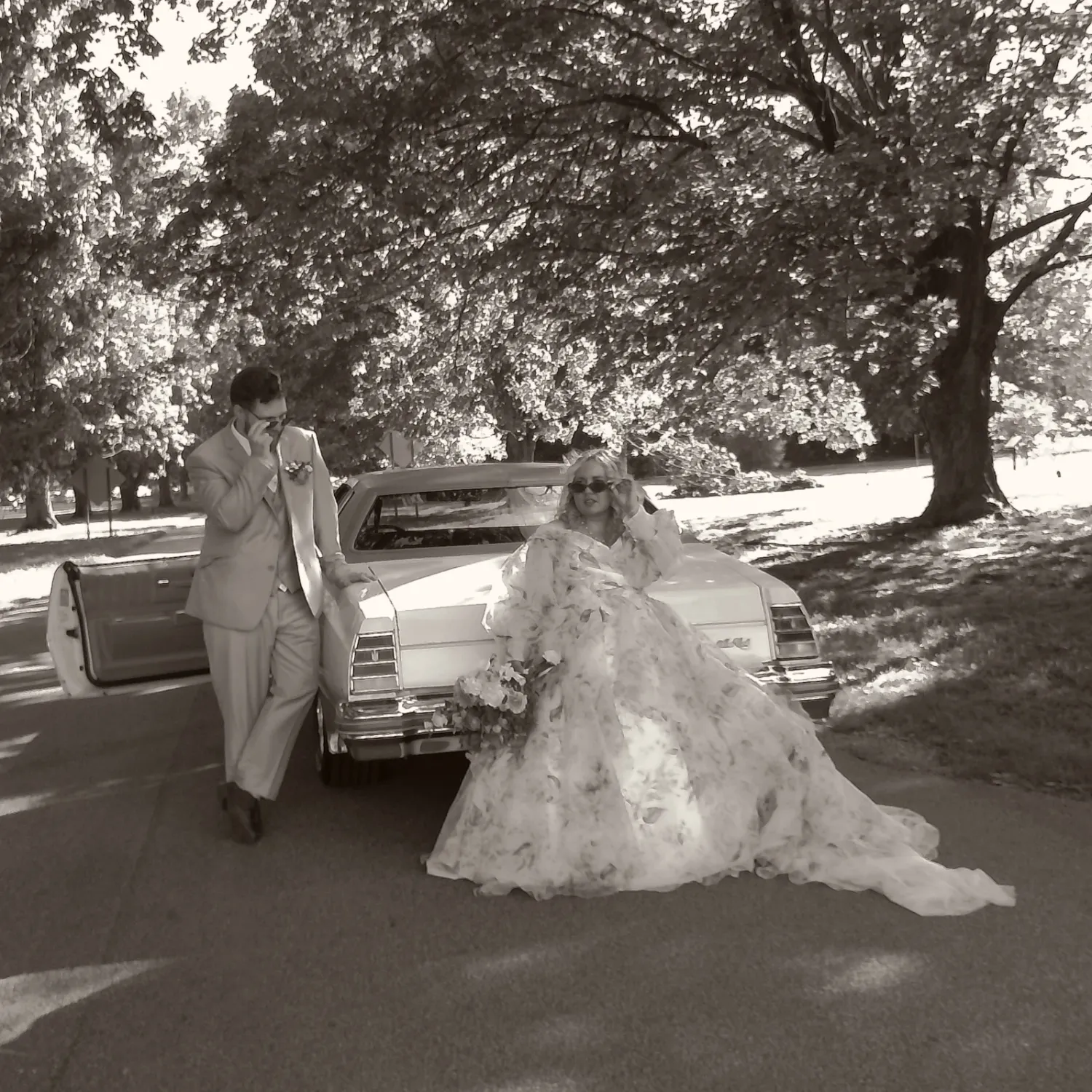 A couple in wedding attire poses near a vintage car in a park setting.