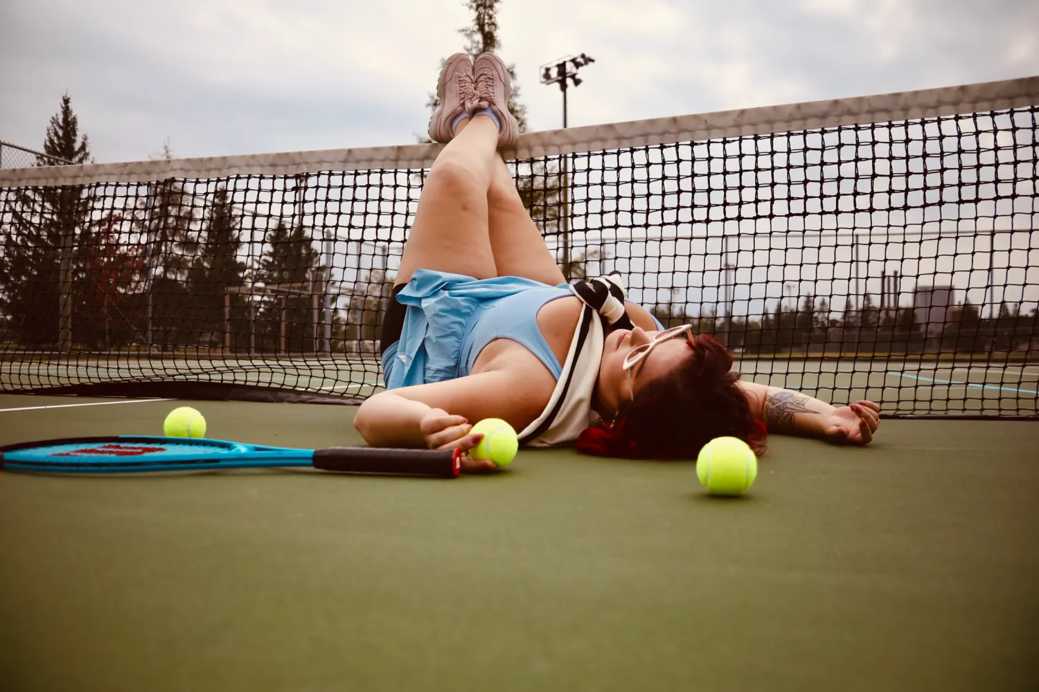 Tennis player lying on court with tennis balls scattered around and net in background.