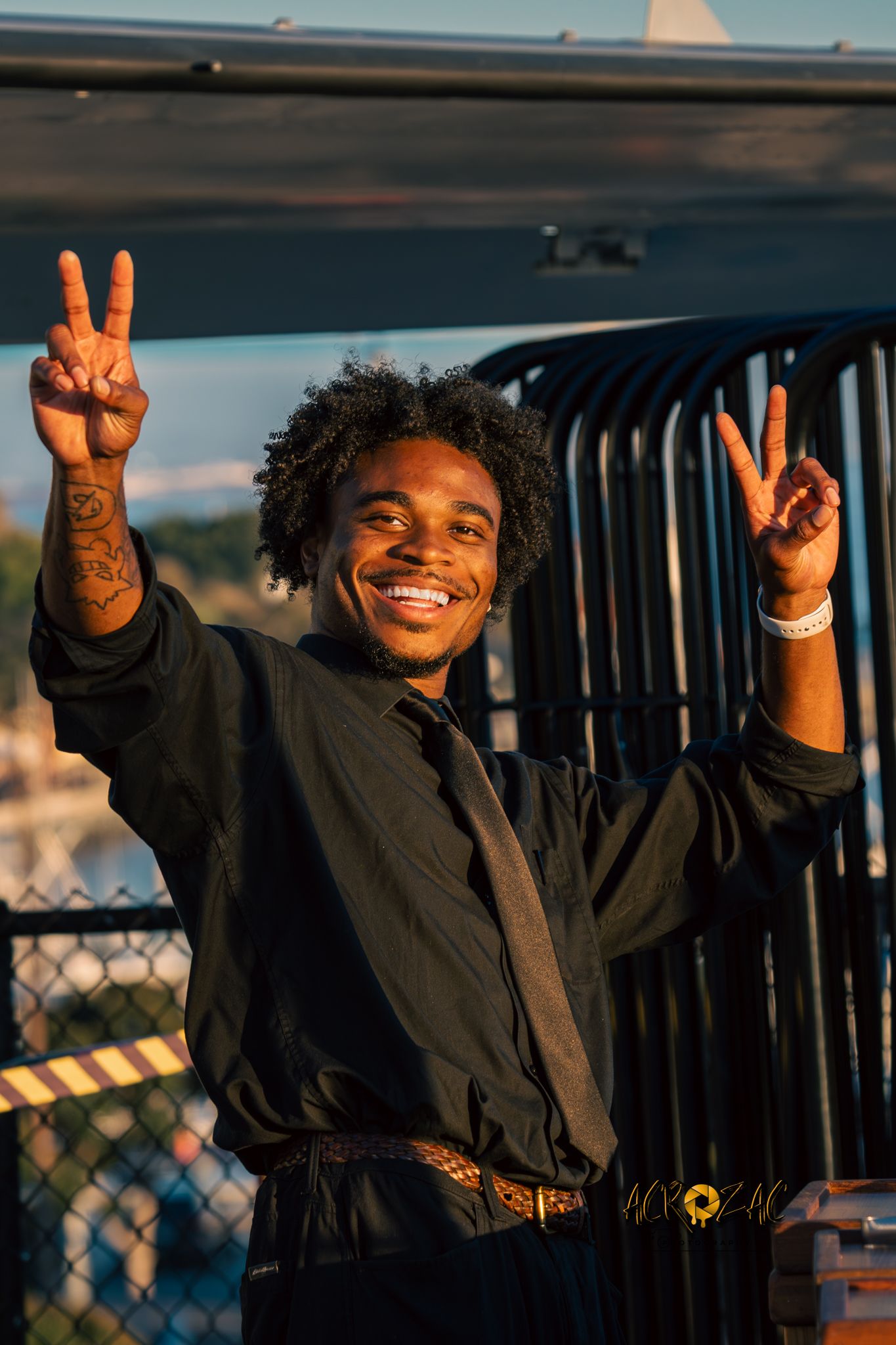 Person in black clothing making peace signs and smiling against sunset backdrop.