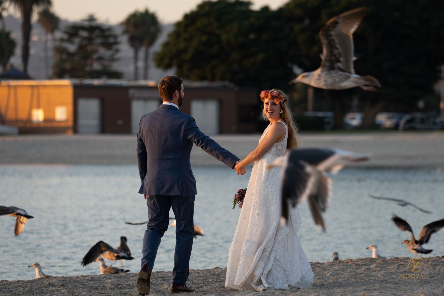Couple walks along waterfront as seagulls fly in the background at sunset.