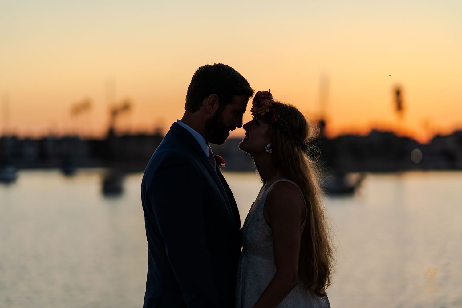 Silhouetted couple against golden sunset sky over calm harbor waters.