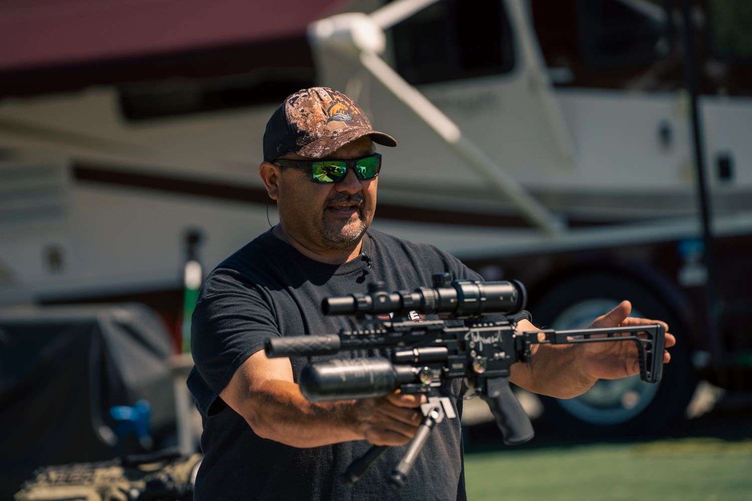 Man in camo cap and sunglasses holding a tactical rifle outdoors near vehicles.