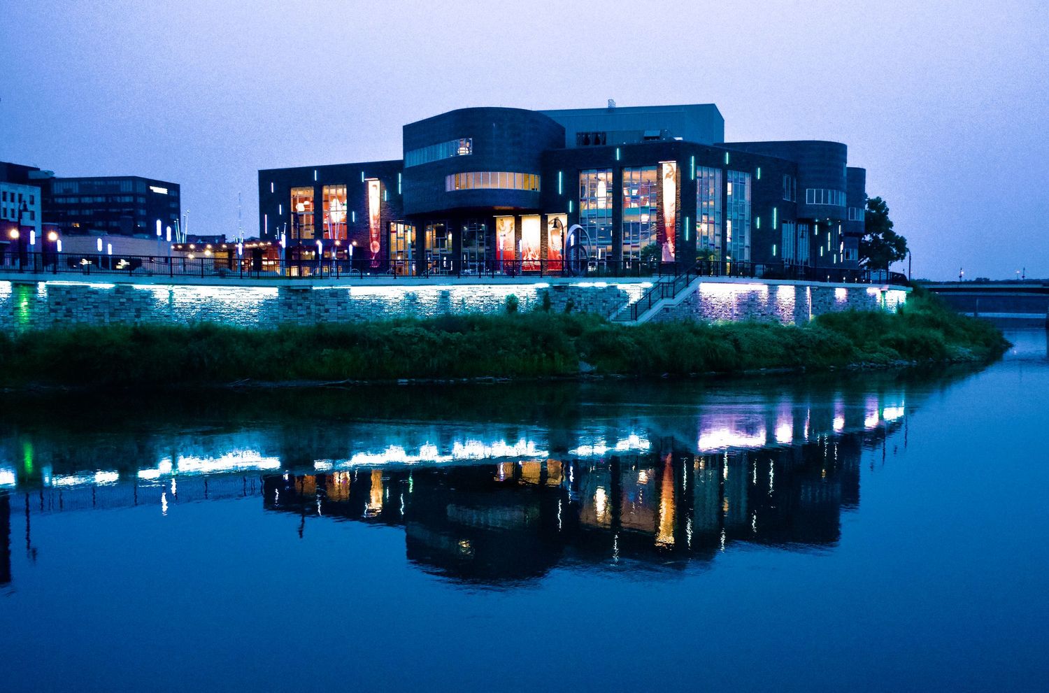 Modern building complex reflecting in calm water at twilight with blue lighting.