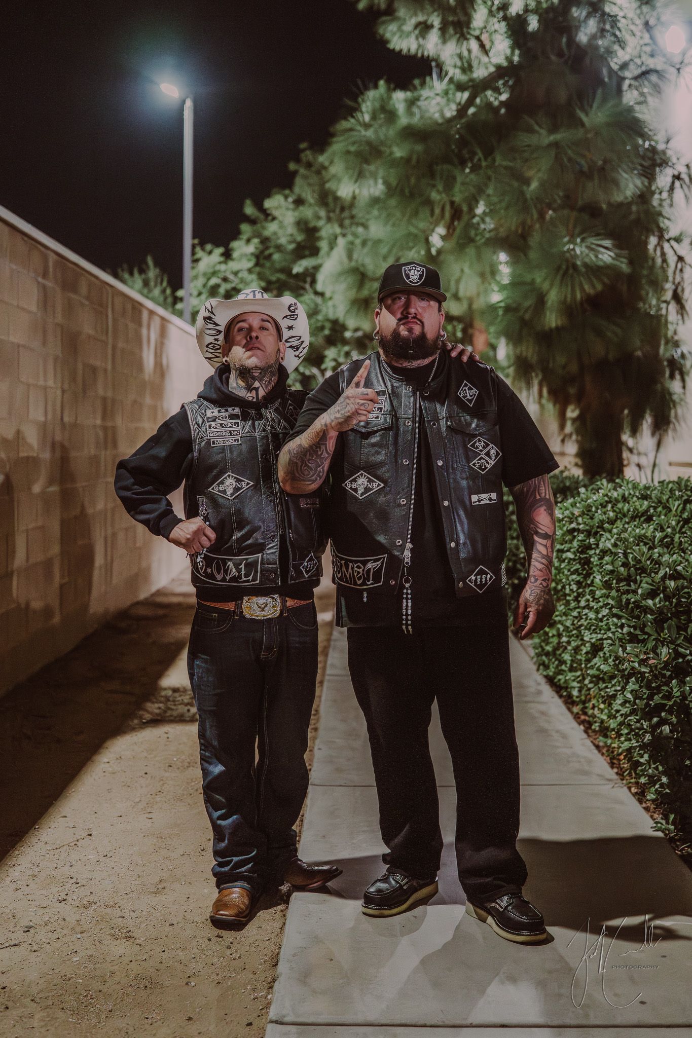 Two men in leather vests standing on sidewalk at night under street lamp.