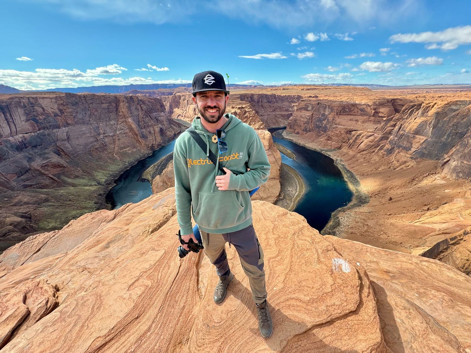 Man smiling at Horseshoe Bend overlook in Arizona, wearing green hoodie, with dramatic canyon and river below.