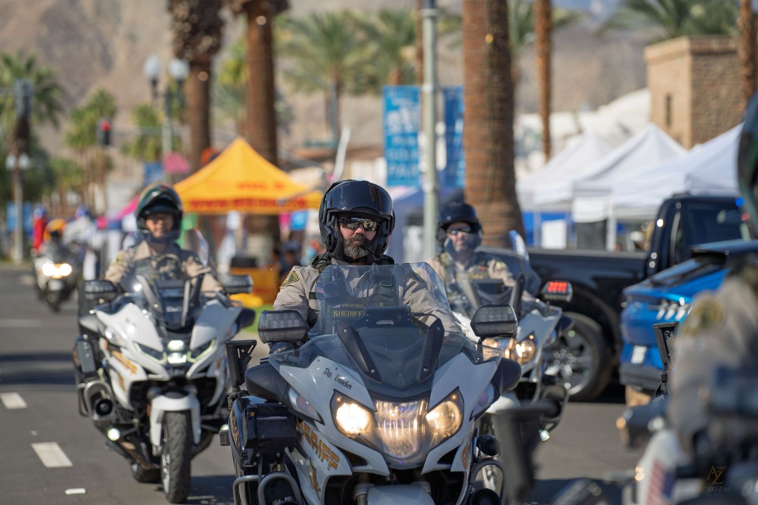 Line of motorcycles and riders parked along a palm tree-lined street.