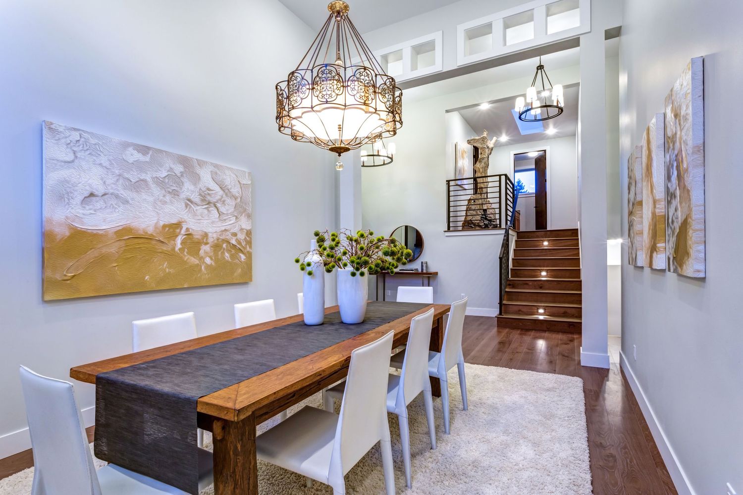 Elegant dining room with wooden table, white chairs, gold chandelier, staircase, and artwork on blue-gray walls.